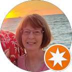 A woman with short brown hair and glasses smiles at the camera, standing by the ocean at sunset. There is an orange star icon in the lower right corner of the image.