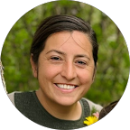 A person with short dark hair smiles at the camera, standing outdoors in front of green foliage and holding a yellow flower.